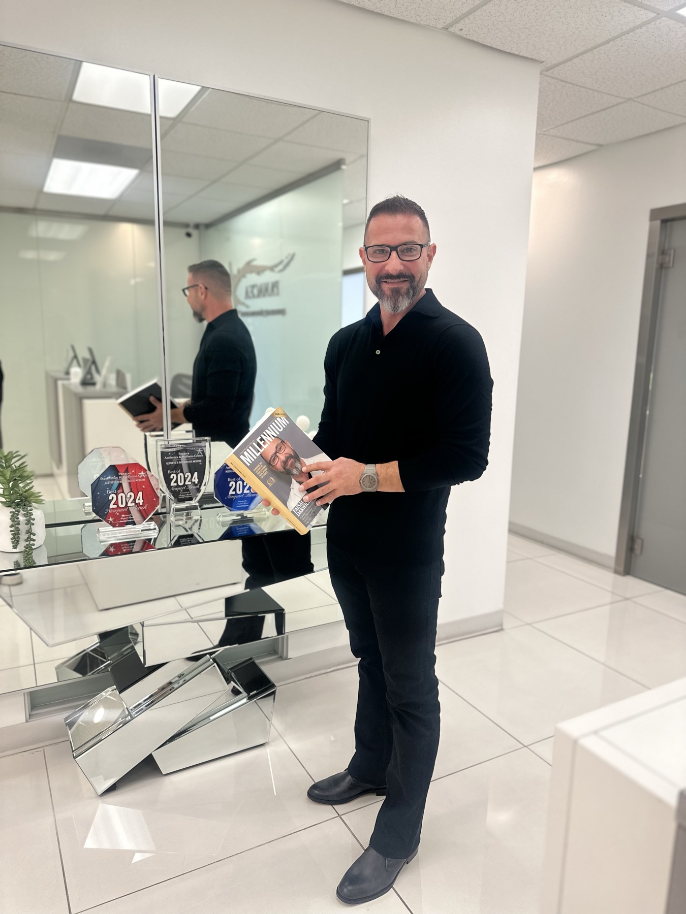 Faisal Darwiche, NP in the Panacea office holding the Millennium magazine feature, beside 2024 award trophies